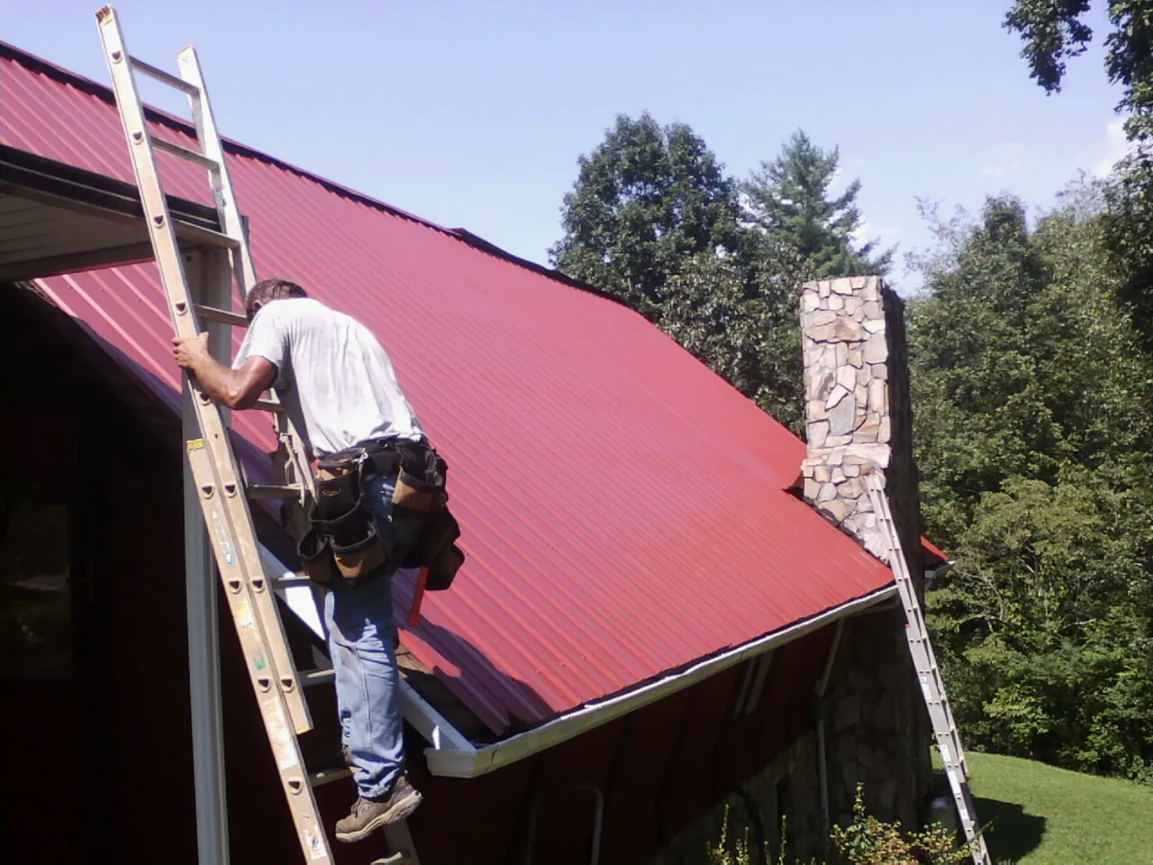 Roofer on a ladder beside a red metal roof and stone chimney — Soffit & Fascia Repair by Roofing Brothers in Luttrell